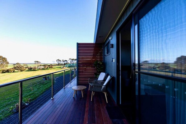 A balcony with a view of a grassy field and a building. There are two chairs and a small table on the balcony.