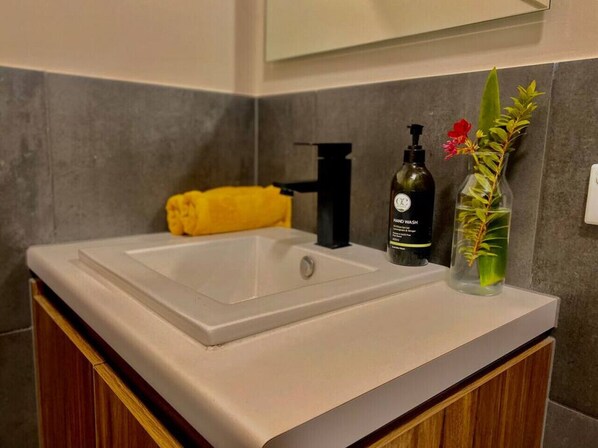 Une salle de bain avec un lavabo blanc, une serviette jaune, un distributeur de savon noir et un vase en verre contenant une plante.