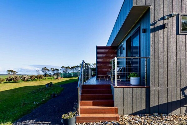 A modern house with a wooden staircase leading to a balcony, a clear view of the surrounding landscape, and a well-maintained lawn.