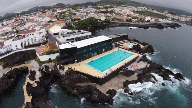 Vista aerea di una località costiera con una piscina ben visibile, circondata da rocce e con una zona residenziale sullo sfondo.