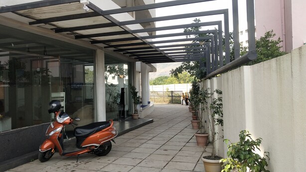 A covered outdoor area with a scooter parked under a pergola, potted plants, and a view of a building in the background. - Hotel Sumanchandra Suites  (Nashik)