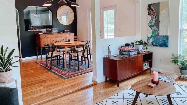 Una sala moderna con una mesa de comedor de madera, un sofá rojo y una mesa de centro. Se ve una cocina al fondo con encimera negra y armarios blancos.