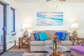 Ein Wohnzimmer mit einem beigefarbenen Sofa, zwei Beistelltischen mit Lampen, einem Glastisch und einem Gemälde von einer Strandansicht.