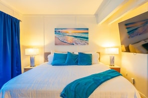 A hotel room with a large bed, two bedside lamps, a painting of a beach scene, and a blue curtain.