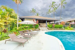 A poolside area with lounge chairs and a covered seating area, surrounded by tropical vegetation and palm trees.