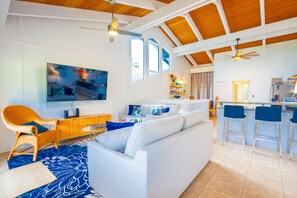 Een ruime woonkamer met een witte bank, een rieten fauteuil, een televisie en een keukenhoek met barstoelen.
