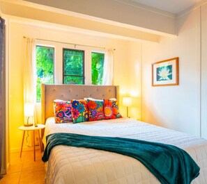 Un dormitorio con cama, almohadas coloridas, cabecera, mesita de noche con lámpara y ventana con cortinas.