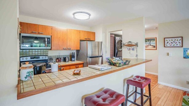 A kitchen with wooden cabinets, a microwave, a refrigerator, and a sink. There are two stools and a small table with a basket.