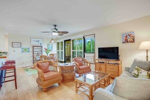A living room with wicker furniture, a ceiling fan, a TV, and a window with shutters.