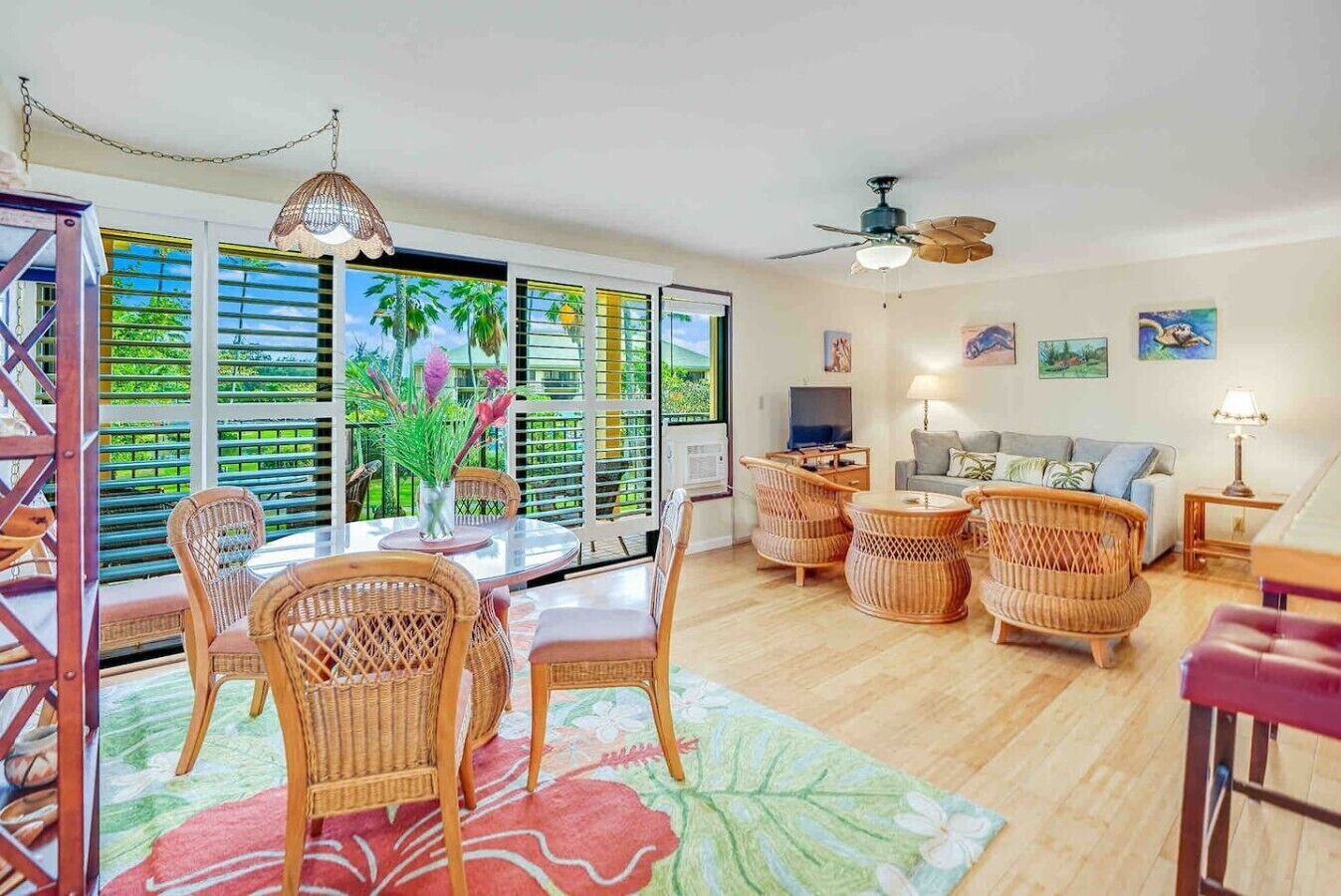A spacious living area with a dining table, wicker furniture, a ceiling fan, and a large window with a view of greenery.