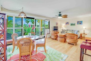 A spacious living area with a dining table, wicker furniture, a ceiling fan, and a large window with a view of greenery.