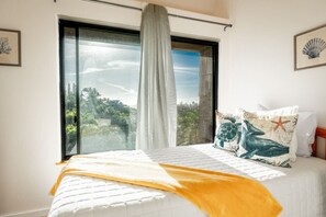 A bedroom with a large window, a bed with a white duvet, a yellow throw blanket, and two decorative pillows with a sea turtle and starfish design.