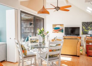 A dining area with a glass table, white chairs, a ceiling fan, and a TV in the background.