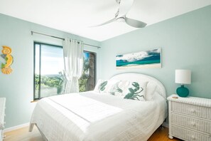 Una camera da letto con un letto, una ventola a soffitto, una finestra con tende, un comodino, una lampada e un quadro appeso al muro.