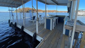 A covered deck area with lounge chairs, a small bar, and a view of a lake with a distant shoreline. - Luxury Main Channel Lake House, Dock, Party Bar (Rocky Mount)