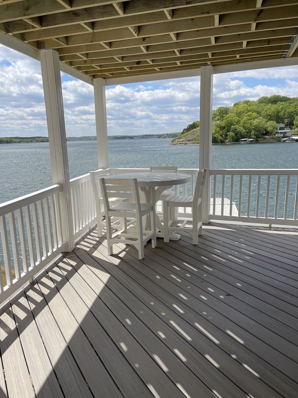 Une terrasse couverte, dotée d’une table et de chaises, qui offre une vue sur une étendue d’eau où l’on aperçoit des bateaux, avec une végétation luxuriante en arrière-plan.