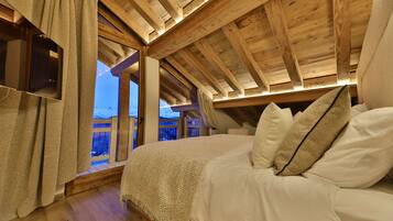 A cozy bedroom with a wooden ceiling, a large bed with white bedding, and a view of a snowy landscape through a window.
