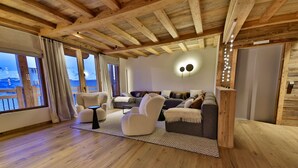 A spacious living room with a wooden ceiling, large windows with curtains, a sofa set, and a wall-mounted light fixture.