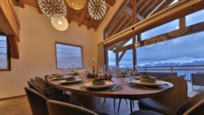 A round dining table set for six with chairs, a view of snow-capped mountains, and a wooden ceiling with hanging lights.