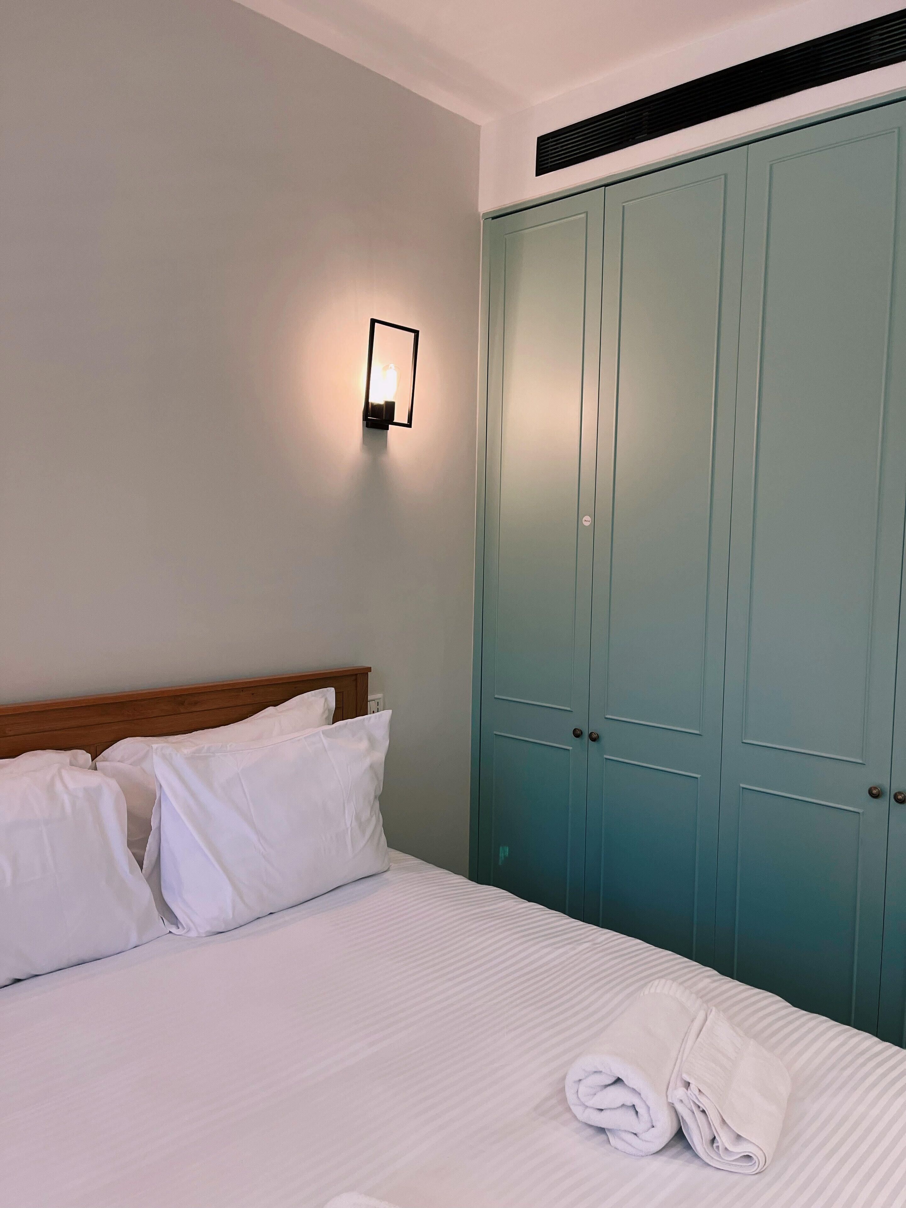 A neatly made bed with white linens and pillows, a wall-mounted light fixture, and a large teal wardrobe.