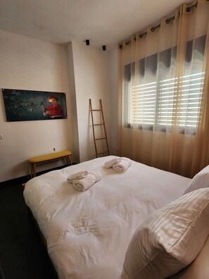 A hotel room with a bed, a bench, a painting, a ladder, and a window with blinds. - Rosa's Place (Jerusalem)