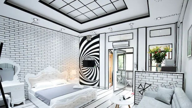 A modern bedroom with a white brick wall, a white bed with a tufted headboard, a white dresser, a white sofa, and a black and white spiral wall design. - Heart of Old Quarter With Balcony View to Street! (ha noi)