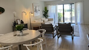 Amplia sala de estar con una mesa de comedor redonda, sillas blancas, un sofá y un ventanal con cortinas.