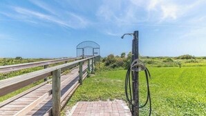 Un muelle de madera que conduce a una reja metálica, con un campo cubierto de césped y un cielo azul despejado al fondo.