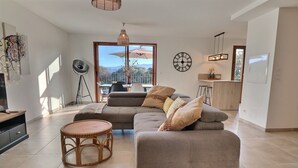 A spacious living room with a grey sofa, a round wicker table, and a dining area with a view of mountains.