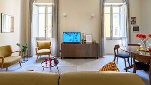 Ein Wohnzimmer mit einer Couch, Sesseln, einem Couchtisch, einem Fernseher auf einer Holzkommode und einem Essbereich mit Tisch und Stühlen.