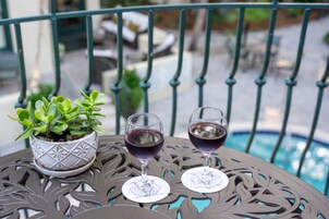 Mesa de metal con dos copas de vino y una planta en maceta en un balcón con vistas a una piscina.