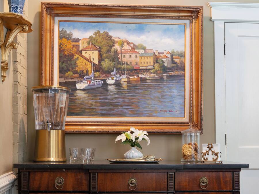 Un dipinto di una cittadina lungo il fiume, un vaso con fiori, un boccale di vetro e un vassoio decorativo su un comò di legno.