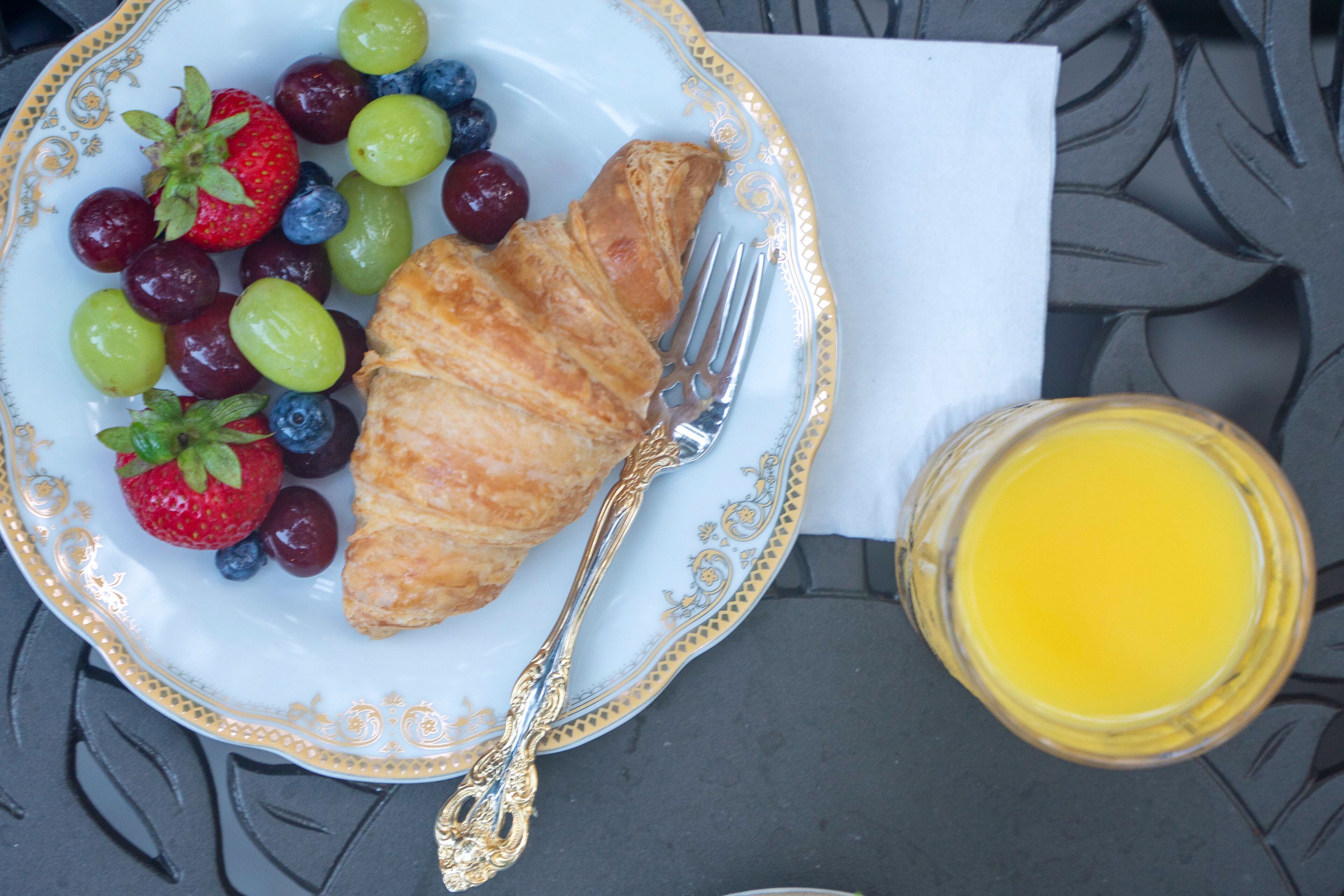 A plate with a croissant, grapes, and strawberries, a fork, and a glass of orange juice on a table.