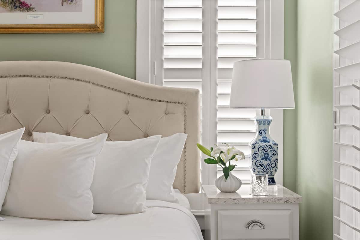 Una cama bien hecha con ropa blanca, un cabecero capitonado y una mesita de noche con un florero y una lámpara.