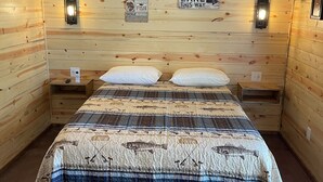 A wooden cabin bedroom with a bed, two wall-mounted lights, a ceiling fan, and a sign that says "LAKE THIS WAY".