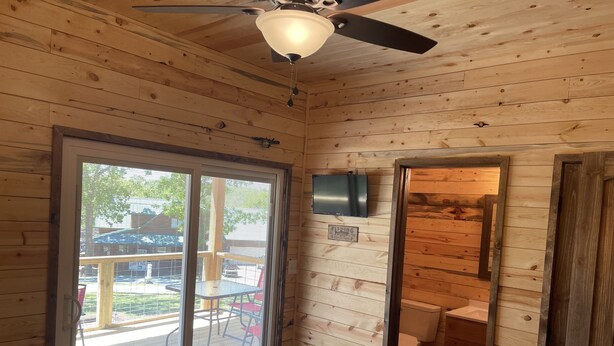 A wooden cabin interior with a sliding glass door to an outdoor deck, a ceiling fan, a toilet, and a television mounted on the wall.