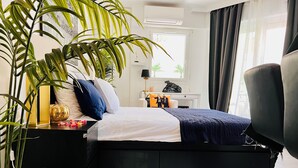 A modern bedroom with a black bed, white pillows, a blue blanket, a large potted plant, a small table with decorative items, and a view of the outside through a window. - 5min Palais, Luxury&Cinema Studio,Croisette,Forville,beaches (Cannes)