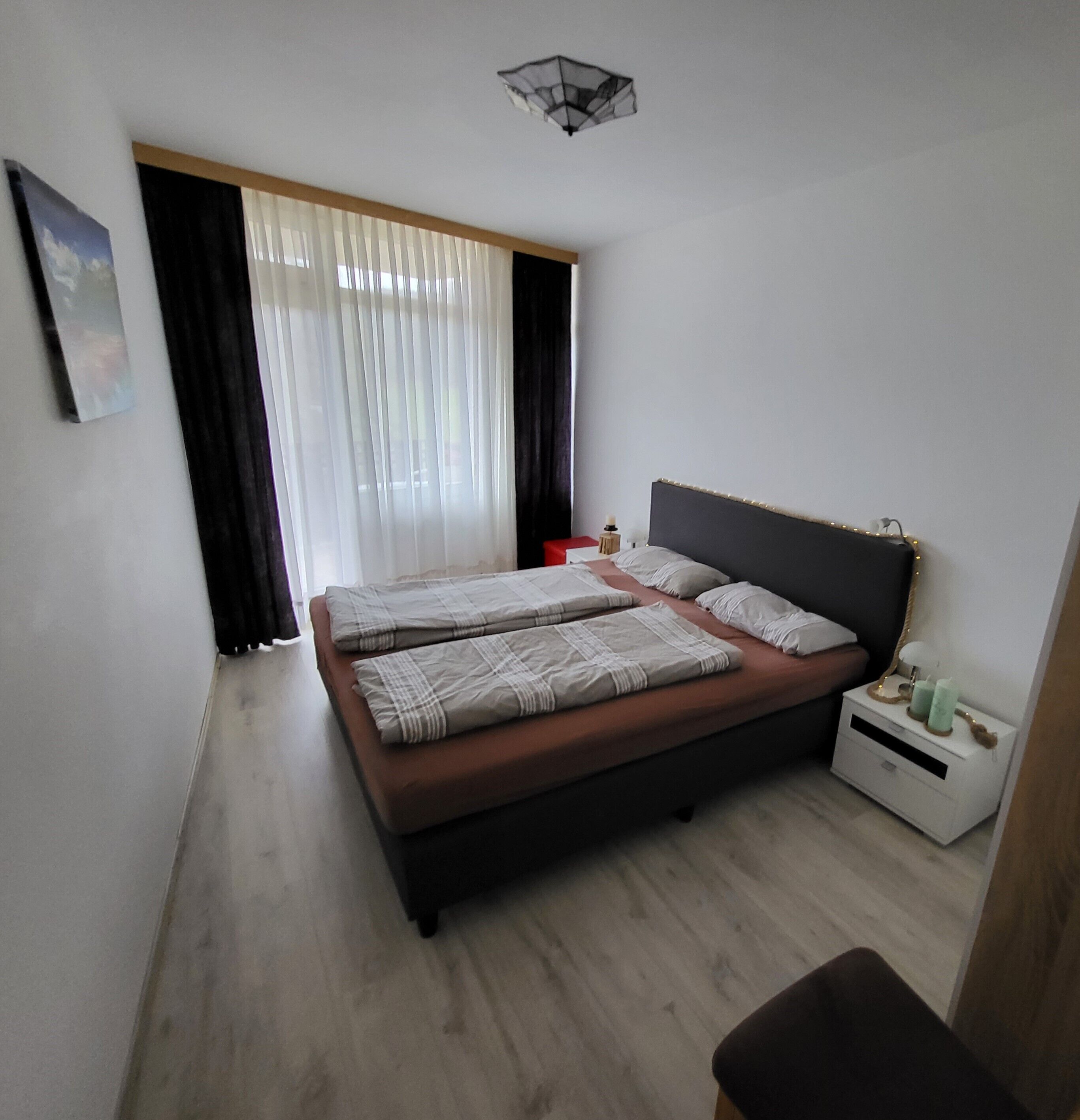Une chambre à coucher avec un grand lit, une table de chevet, une commode, une fenêtre avec des rideaux et un luminaire au plafond.