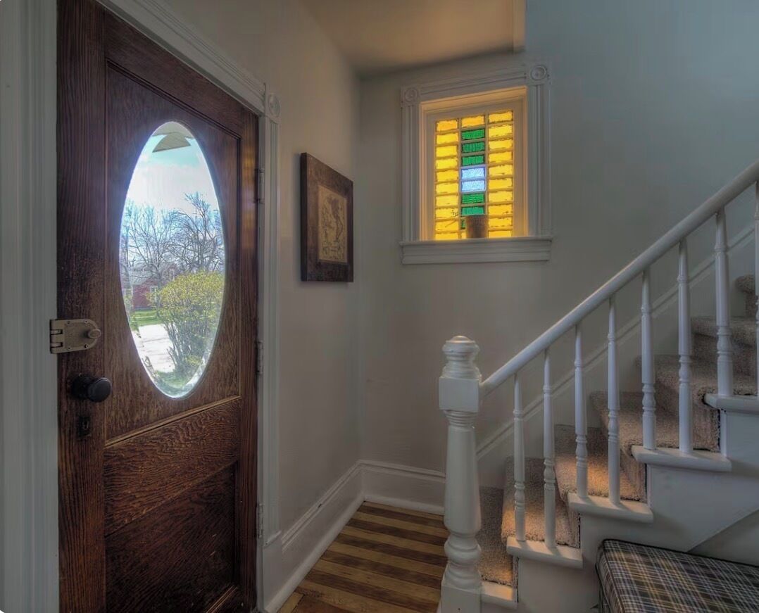 Une porte en bois avec un panneau de verre, un escalier blanc avec une rampe et une petite fenêtre ornée de vitraux colorés.