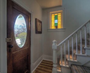 Una puerta de madera con un panel de vidrio, una escalera blanca con barandilla y un ventanal con vidrios de colores.