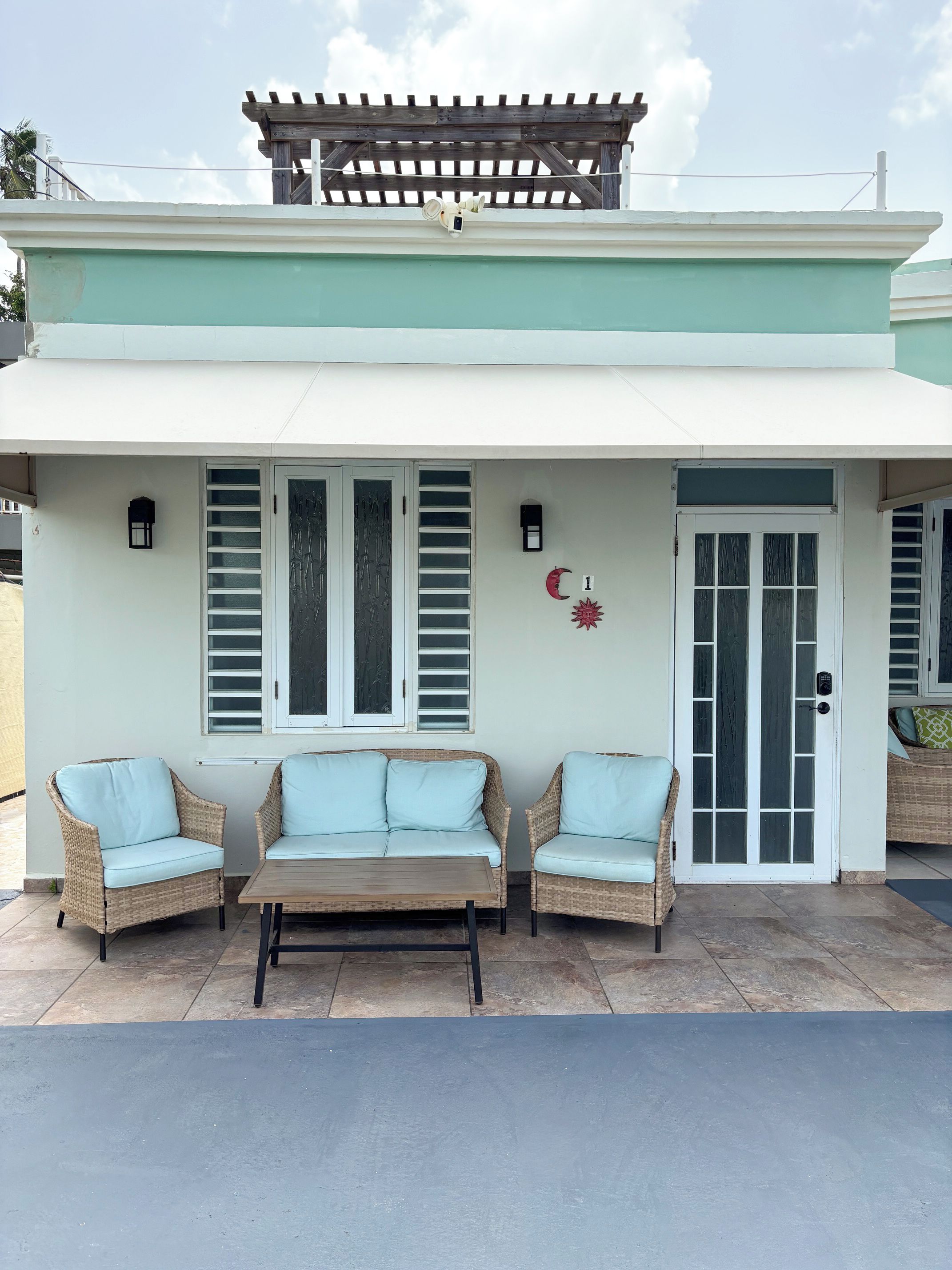 A building with a covered porch, two wicker sofas with light cushions, a wooden table, and a pergola.