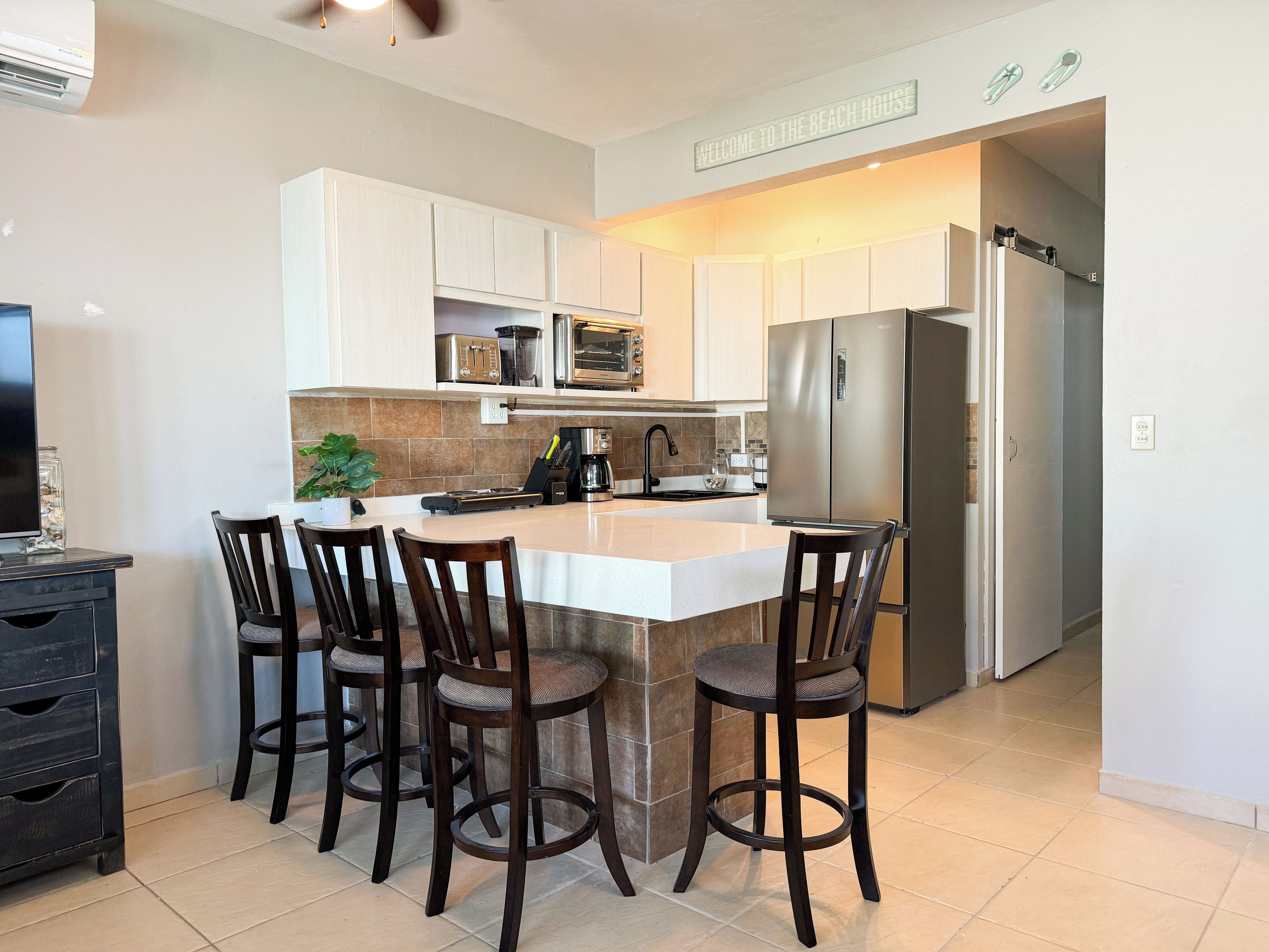 A modern kitchen with a central island, bar stools, a refrigerator, and a sign that reads 'WELCOME TO THE BEACH HOUSE'.