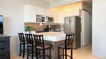 A modern kitchen with a central island, bar stools, a refrigerator, and a sign that reads 'WELCOME TO THE BEACH HOUSE'.