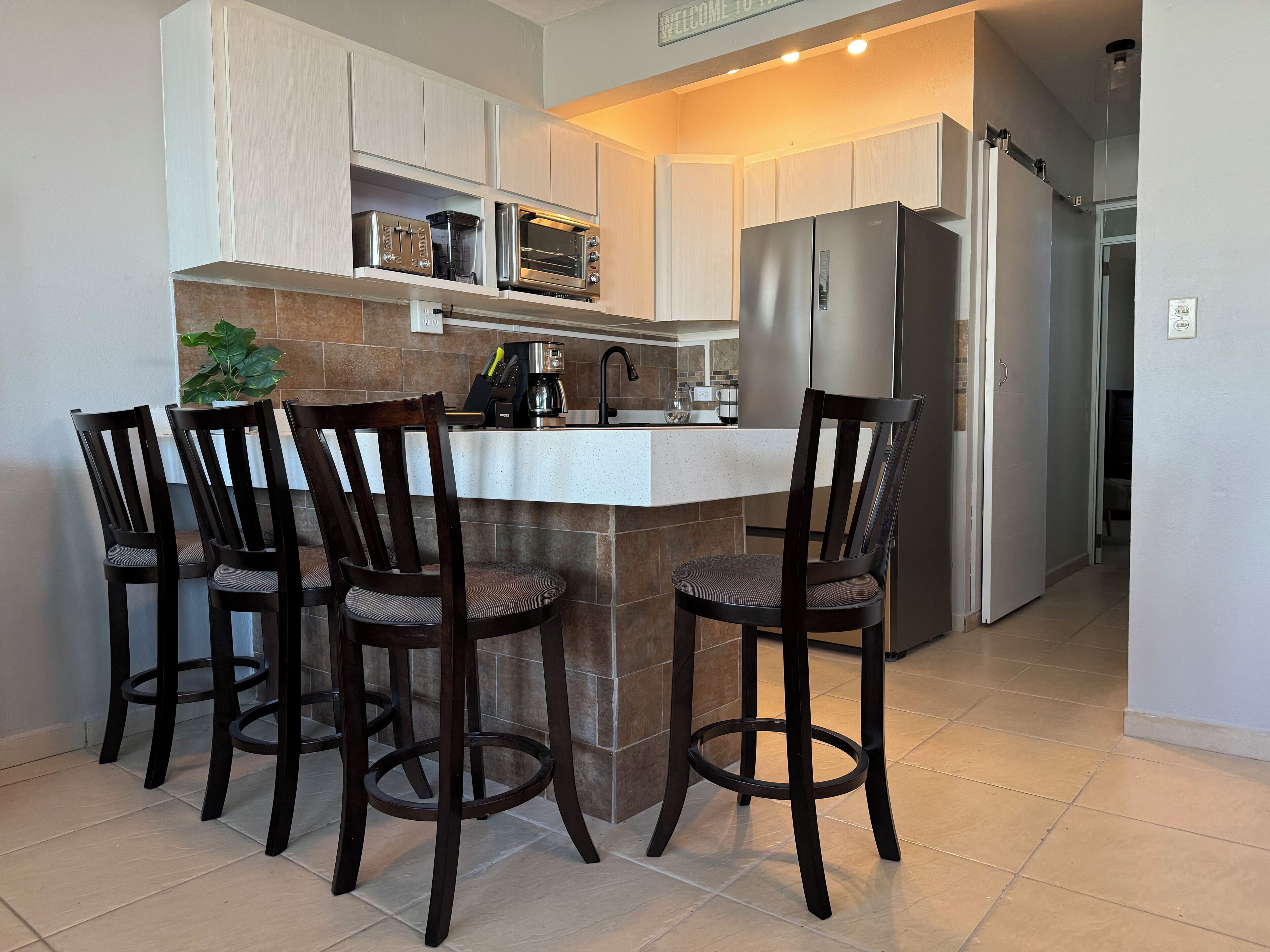 A kitchen with a central island, four bar stools, a refrigerator, and a coffee maker.