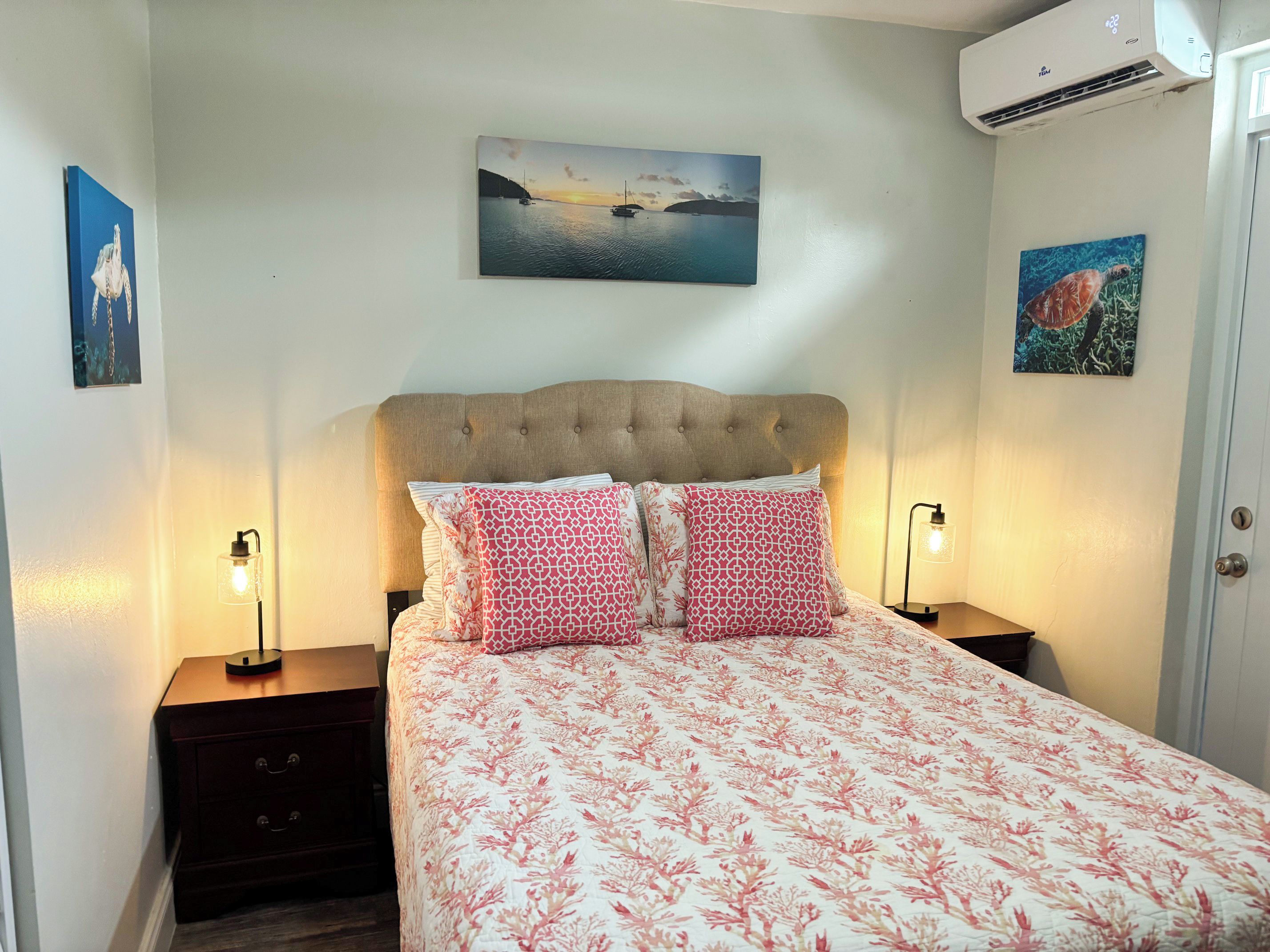 Une chambre à coucher comprenant un lit, deux tables de chevet, un climatiseur fixé au mur et deux tableaux encadrés.