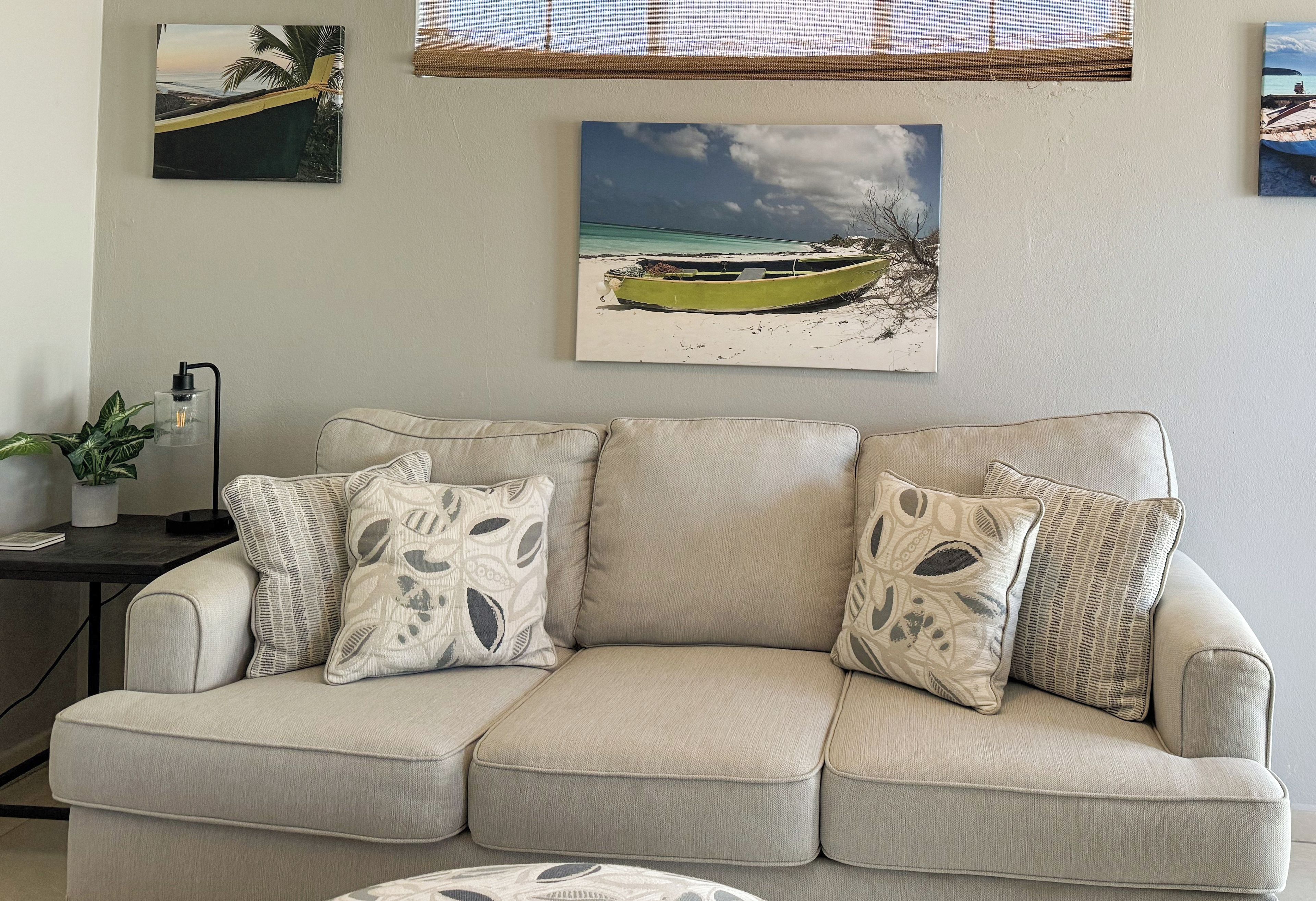 Ein beiges Ecksofa mit gemusterten Kissen, ein kleiner Beistelltisch mit einem Blumentopf und Wandbilder, die eine Strandlandschaft zeigen.
