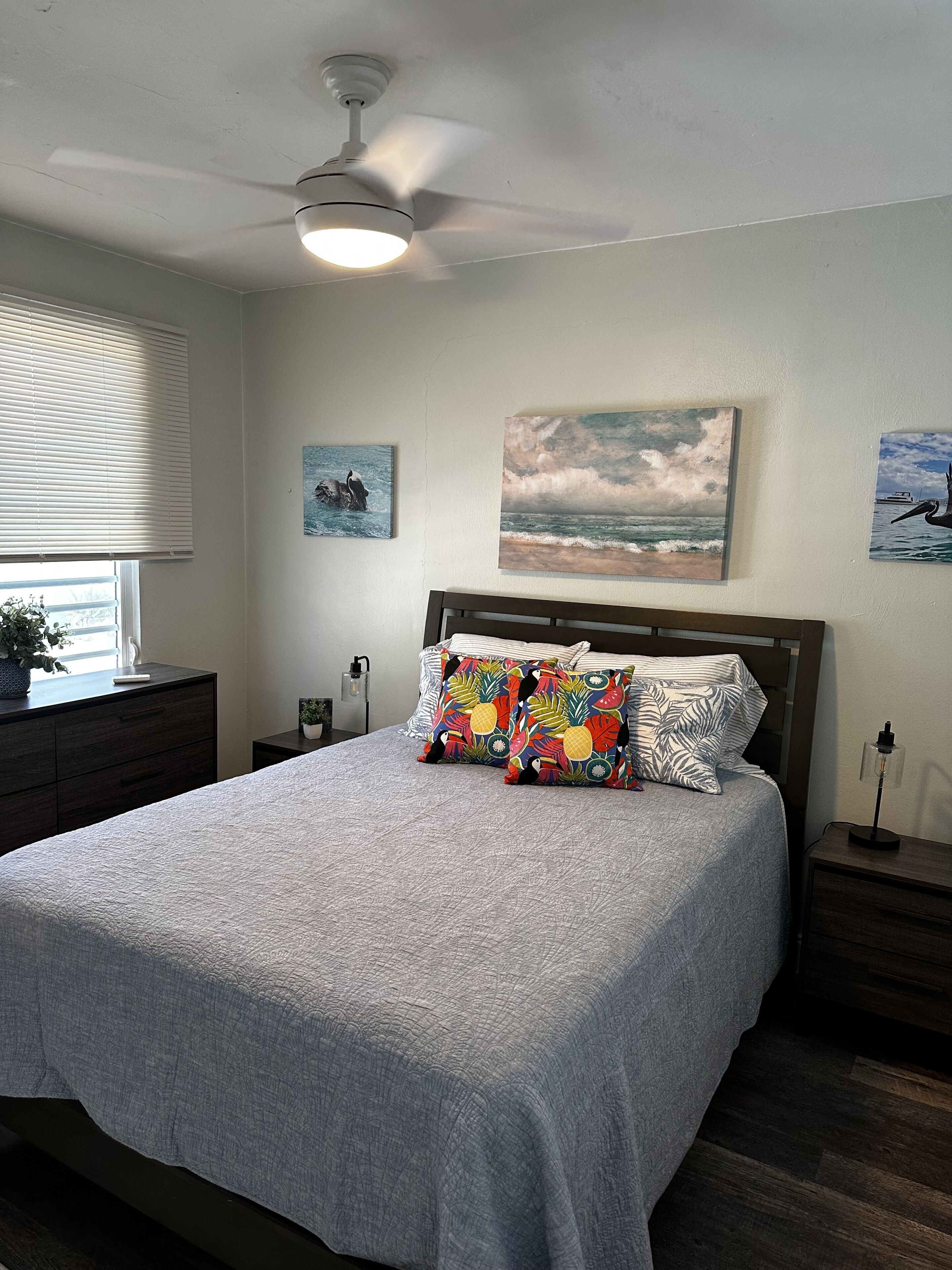 Une chambre à coucher comprenant un lit, un ventilateur de plafond, une table de chevet et une commode, ainsi qu’une fenêtre avec des stores.