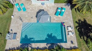 An aerial view of a rectangular swimming pool with a tiled deck surrounding it, equipped with lounge chairs and umbrellas.