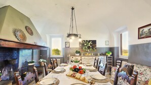 Un comedor con una larga mesa dispuesta para seis personas, rodeada de sillas de madera. Un candelabro cuelga del techo, y la habitación cuenta con una chimenea, una mesa de comedor con una bandeja de frutas y quesos, y un sofá floral.