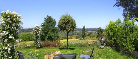 A garden with two metal chairs and a table, surrounded by potted plants and a view of the countryside.